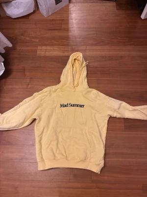 Gul hoodie  Mad Summer - Snygg gul hoodie med svart broderad text på bröstet. Klassisk känguruficka framtill och huva med dragsko. Tillverkad i mjukt material som känns skönt mot huden. Perfekt för dig som gillar streetwear och vill sticka ut lite extra. Det är för herrar men funkar också på kvinnor