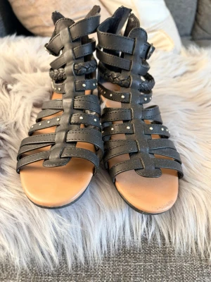 Svarta gladiatorsandaler med nitar - Snygga svarta gladiatorsandaler med flätade detaljer och små silverfärgade nitar. Sandalerna har flera remmar i konstläder och dragkedja bak för enkel på- och avtagning.