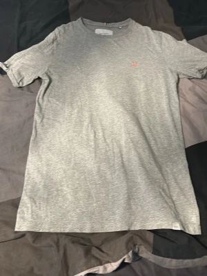 Grå t-shirt från Les Deux - Snygg gråmelerad t-shirt från Les Deux med liten orange broderad logga på bröstet. Klassisk rund halsringning och korta ärmar. Tillverkad i mjuk bomull som känns skön mot huden. Perfekt basplagg för alla tillfällen.