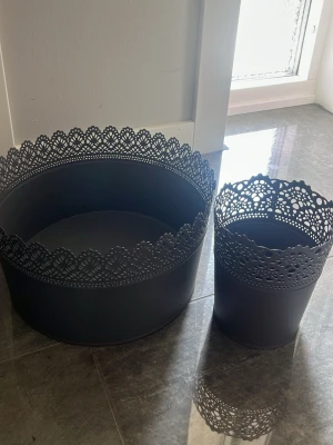 Svarta metallkorgar med spetskant - Två svarta runda metallkorgar med dekorativ spetskant upptill. Perfekta för förvaring eller som inredningsdetalj. Den ena är större och den andra mindre, båda har ett elegant och stilrent utseende.