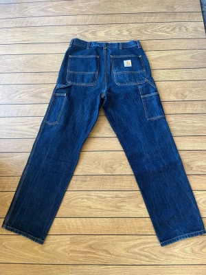 Carhartt Single knee - Säljer ett par mörkblå single knee jeans från Carhartt med raka ben och flera praktiska fickor. Klassisk utility-stil med kontrastsömmar och Carhartt-logga på bakfickan. Tillverkade i slitstark denim, perfekta för en workwear streetwear-look. Endast testade, som nya!!!