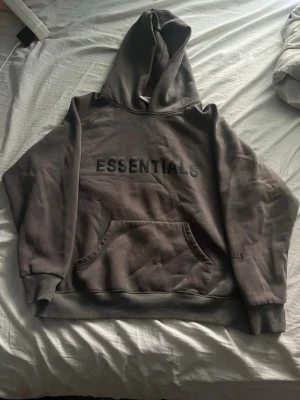 Brun Essentials hoodie med huva - Säljer en brun hoodie från Essentials med stor huva och magficka. Hoodien har tryckt logga på bröstet och diskret text på ärmen. Tillverkad i mjukt bomullsmaterial och har ribbade muddar vid ärmslut och nederkant. Perfekt för en avslappnad och trendig look.