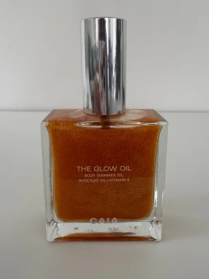 The Glow Oil Body Shimmer från CAIA - Oanvänd  - The Glow Oil Body Shimmer från CAIA ger huden en skimrande finish med guldigt skimmer. Innehåller avokadoolja och vitamin E för extra vård. Kommer i en fyrkantig glasflaska med silverfärgat lock och har en lyxig, oljig konsistens.