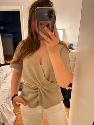 Beige omlottopp från Zara - Trendig beige omlottopp från Zara med korta ärmar och djup V-ringning. Toppen har en snygg twistdetalj framtill som ger en cool siluett. Perfekt att styla med både jeans och kjol.