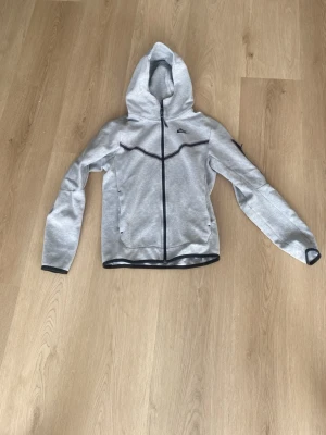 Grå Nike hoodie med dragkedja - Snygg grå hoodie från Nike med hel dragkedja framtill och svart detalj längs bröstet. Tröjan har huva, två fickor och svarta kantband vid ärmslut och nederkant. Perfekt för en sportig och avslappnad stil.