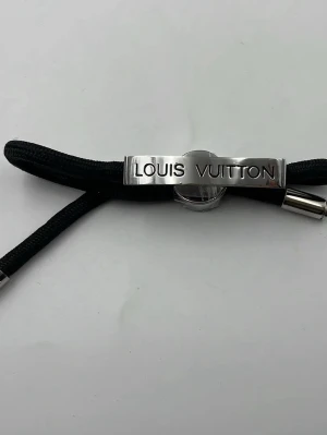Louis Vuitton svart armband - Snyggt svart armband från Louis Vuitton med silverfärgat metallspänne där märkesnamnet är ingraverat. Armbandet har en stilren och modern design med justerbar längd och kommer med originalförpackning och dustbag.