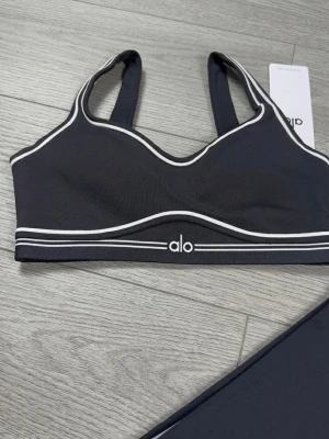 Alo Yoga - Snyggt set från Alo med svart sport-bh och matchande tights. Båda plaggen har vita kontrastlinjer och Alo-logga framtill. Materialet är stretchigt och följsamt, perfekt för träning eller chill. Sport-bhn har bred resår och tightsen har hög midja.