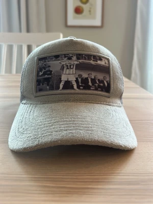 Beige Il Capo keps med fotbollsmotiv - Säljer en beige keps från Il Capo med mesh på baksidan och ett svartvitt fotomotiv framtill som visar en fotbollsspelaren Messi med nummer 10. Kepsen har böjd skärm och justerbar passform. Materialet är en mix av tyg och mesh för extra ventilation.
