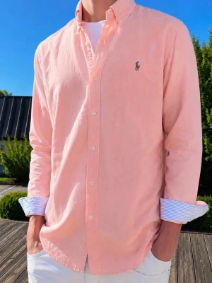 Ljusrosa skjorta från Ralph Lauren - FÖRGEN ÄR KORRAL/ROSA (Moddelen är 185 och väger 76KG)Säljer en stilren ljusrosa skjorta från Ralph Lauren med klassisk button-down krage och broderad logga på bröstet. Skjortan har långa ärmar och vita knappar, perfekt för en clean och fräsch look.