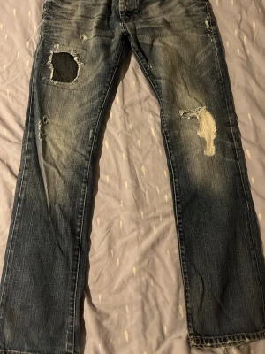 Blå slitna jeans med patchade hål - Ett par blå jeans med raka ben och tydliga slitningar samt patchade hål på båda benen. Jeansen har en tvättad look och synliga lagningar som ger en cool, avslappnad stil. Perfekta för dig som gillar en edgy och trendig vibe.