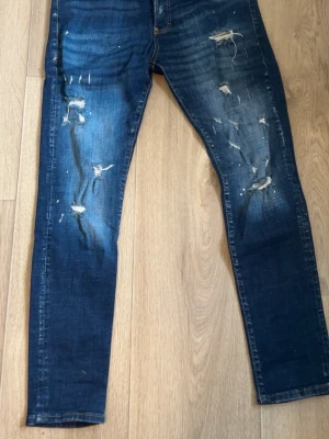 Mörkblå slitna skinny jeans - Säljer ett par mörkblå skinny jeans med slitningar och distressed detaljer på både fram- och baksida. Jeansen har en tight passform och coola slitna partier som ger en trendig look. Perfekta för dig som gillar streetstyle och vill ha en edgy vibe.