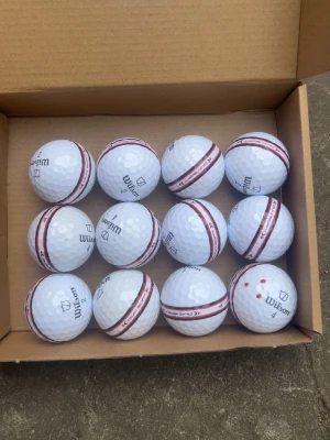 Wilson Staff stripe 12-pack - Säljer ett dussin vita golfbollar från Wilson med en tydlig röd och svart rand runt om. Bollarna har klassisk dimple-yta och märkning med Wilson-logga och siffror. Perfekta för dig som vill ha stilrena och lättigenkännliga golfbollar på banan.