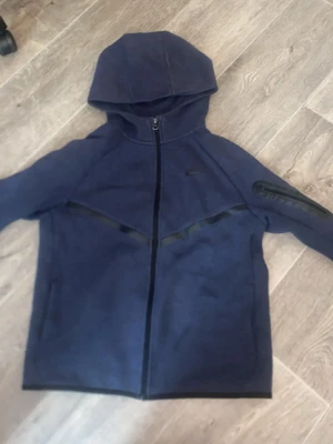 Mörkblå Nike hoodie med dragkedja - Snygg mörkblå hoodie från Nike med hel dragkedja framtill och huva. Hoodien har svarta detaljer längs bröstet och en ficka med dragkedja på ena ärmen. Tillverkad i mjukt material som passar perfekt till en avslappnad stil.