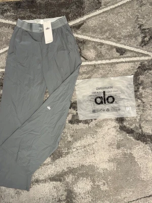 Grå mjukisbyxor från Alo - Snygga grå mjukisbyxor från Alo med elastisk midja och diskret logga på benet. Tillverkade i ett mjukt och stretchigt material som känns skönt mot huden. Perfekta för chill eller träning. Klassisk rak passform och stilren look.