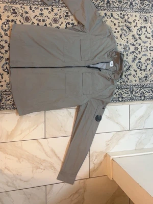 Beige vindjacka från C.P. Company - Stilren beige vindjacka från C.P. Company med huva, två bröstfickor och dragkedja framtill. Jackan har en ikonisk linsdetalj på ärmen och meshfoder på insidan. Perfekt för en urban och sportig look.
