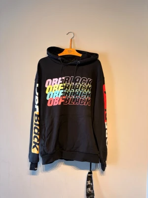 Svart hoodie med färgglad OBF-tryck - Säljer en svart hoodie med stor färgglad OBF Black-tryck på bröstet i rosa, gul, blå och röd. Tryck även på båda ärmarna. Hoodien har huva med dragsko och en stor magficka. Perfekt för dig som gillar streetwear och vill sticka ut.