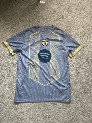 blå Barcelona matchtröja - Snygg grå Barcelona matchtröja med gula detaljer och klubbmärke på bröstet. Tröjan har korta ärmar, rund hals och ett stort Spotify-tryck på magen. Tillverkad i ett lätt och ventilerande material som passar perfekt till träning eller match. passar som xl