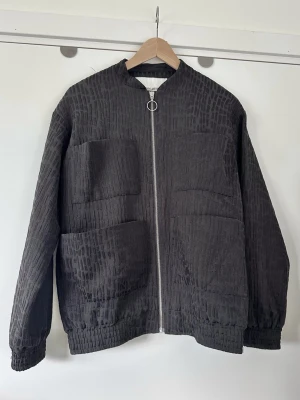 Carin Wester - Svart bomberjacka med struktur - storlek XS / S - Stilren svart tunn oversize bomberjacka / jacka från Carin Wester med en unik texturerad yta. Storlek XS, men är oversized så passar även S, men är då lite mer figurnära. Tidlös modell med dragkedja framtill med en ringformad dragtapp samt två bröstfickor och två större fickor nedtill. Muddar finns vid ärmslut och nederkant. Tidlös klassiker som passar till allt. Mycket gott skick, som ny!