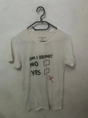 Vit t-shirt med rolig text - Vit t-shirt från Legend med trycket 'AM I DRUNK? NO YES' och en röd bock utanför rutorna. T-shirten har rund hals och korta ärmar. Perfekt för dig som gillar humoristiska plagg och vill sticka ut lite extra.
