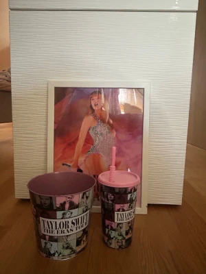 Taylor Swift The Eras Tour merch set - Säljer ett set med Taylor Swift The Eras Tour merch: en poster i vit ram, en popcornhink och en dryckesmugg med sugrör. Både hinken och muggen har collage med bilder på Taylor Swift i rosa, svart och vitt. Perfekt för fans och samlare!