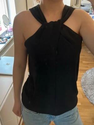 Svart halterneck topp från Zara - Säljer en svart halterneck topp från Zara med snygg twistad detalj vid halsen. Toppen är ärmlös och har en lös passform som ger en avslappnad men ändå stilren look. 