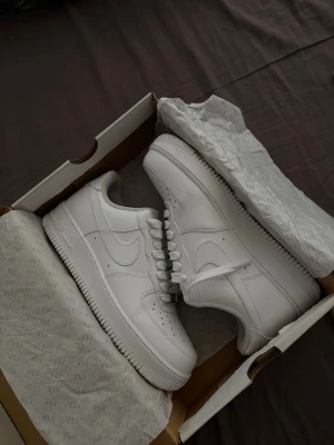Vita Nike Air Force 1 sneakers - Klassiska helvita Nike Air Force 1 sneakers i lågt utförande. Skorna har en stilren design med perforerad tå, vit sula och ikoniska swoosh-loggan på sidan. Tillverkade i slätt läder med vita skosnören och diskreta detaljer. Perfekta för en clean och tidlös look.