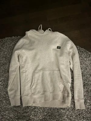 Ljusgrå hoodie från Les Deux - Säljer en stilren ljusgrå hoodie med huva och dragsnören. Tröjan har en stor magficka framtill och en liten svart patch med siffran 'II' på bröstet. Tillverkad i mjukt bomullsmaterial, perfekt för en avslappnad look.
