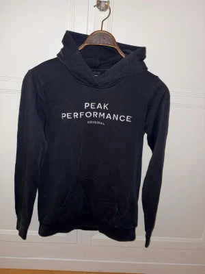 Mörkblå Peak Performance Hoodie - Klassisk modell med huva och känguruficka framtill. Tillverkad i mjukt bomullsmaterial och har ribbade muddar vid ärmslut och nederkant. Perfekt för en avslappnad och sportig stil. Passar för dig med storlek 164-XS