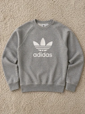 Grå sweatshirt från Adidas - Klassisk grå sweatshirt från Adidas med stort vitt Trefoil-logo tryck på bröstet. Tröjan har rund halsringning, långa ärmar och ribbade muddar vid ärmslut och nederkant. Perfekt för en avslappnad och sportig stil.