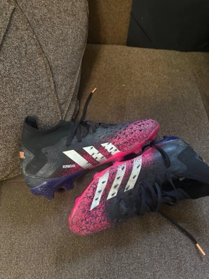 Adidas fotbollsskor - Säljer ett par snygga Adidas fotbollsskor med svart och rosa färgskiftning, vita ränder på sidorna och hög ankelkrage. Skorna har snörning och mönstrad ovandel för extra grepp på planen. Perfekta för dig som vill sticka ut på fotbollsplanen.