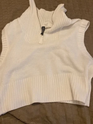 Vit stickad väst med halv dragkedja - Snygg vit stickad väst med hög krage och halv dragkedja framtill. Västen har bred ribbad kant nedtill och vid ärmöppningarna, vilket ger en trendig och avslappnad look. Perfekt att styla över en skjorta eller t-shirt.