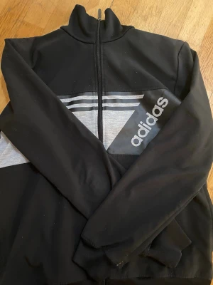 Svart och grå zipjacka från Adidas - Svart jacka med hel dragkedja från Adidas, har gråa och svarta ränder över bröstet och Adidas-logga på ena sidan. Perfekt för träning eller chill dagar.(jackan är helt ny det vita är bara fläckar,det går bort lätt) 