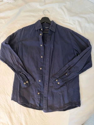 Blå linne skjorta från Jack & Jones - Säljer en stilren blå skjorta från Jack & Jones med klassisk krage och knappar framtill. Skjortan har långa ärmar och är tillverkad i mjuk bomull, perfekt för en avslappnad men snygg look.
