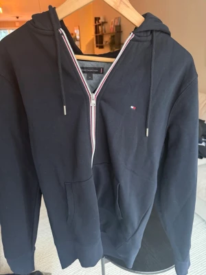 Svart hoodie från Tommy Hilfiger - Snygg svart hoodie från Tommy Hilfiger med dragkedja och klassiska röda, vita och blå detaljer längs dragkedjan. Har huva med dragsko, fickor framtill och liten broderad logga på bröstet. Perfekt för en chill och stilren look.