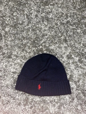 Mörkblå ribbstickad mössa Polo - Snygg mörkblå ribbstickad mössa från Polo Ralph Lauren med uppvikt kant och klassisk röd logobrodyr framtill. Perfekt accessoar för kyliga dagar och enkel att matcha med olika outfits.