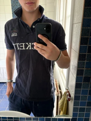 Marinblå Hackett pike - Snygg marinblå polo t-shirt från Hackett med tryck och broderi framtill, nummerdetalj och kontrastkrage i vinrött. Kortärmad modell med blåa och vinröda detaljer på ärmslut och krage. Perfekt för dig som gillar sportig stil. Liten fläck på baksidan av armen och på framsidan av piken, går nog att få bort.