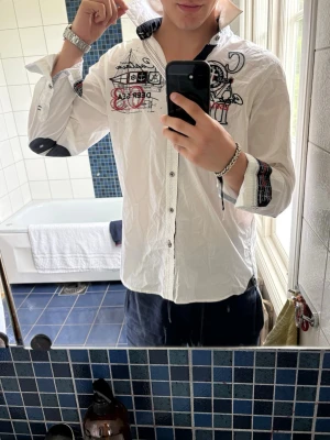Vit skjorta från Camp David med broderi - Säljer en vit skjorta från Camp David med stora broderade detaljer och tryck på bröstet och ärmen. Skjortan har klassisk krage, knappar framtill och svarta kontrastdetaljer på insidan av kragen. Perfekt för dig som gillar statement-plagg.