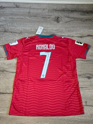 Portugal matchtröja Ronaldo 2026 - Röd Portugal matchtröja från Puma med nummer 7 och texten 'Ronaldo' på ryggen. Tröjan har korta ärmar, vågigt mönster, gröna detaljer vid krage och ärmslut samt broderad landslagssköld. Tillverkad i lätt funktionsmaterial.