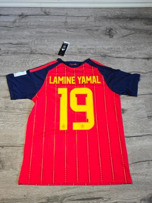 Lamine Yamal jersey 2026 - Snygg röd matchtröja från Adidas för Spaniens landslag med gula detaljer och nummer 19. Tröjan har blå ärmar med gula ränder på axlarna och namntryck 'LAMINE YAMAL' på ryggen. Perfekt för fotbollsfans som vill stötta sitt lag.