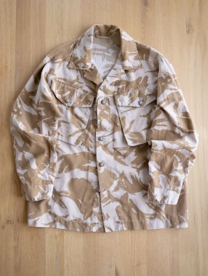 Beige Army Jacket, size L - Overshirt i beige och vitt camomönster med två bröstfickor och knappar framtill. Skjortan har klassisk krage och raka ärmar. Tillverkad i ett kraftigare bomullstyg som ger en robust känsla. Perfekt för lager på lager och streetwear-stil.