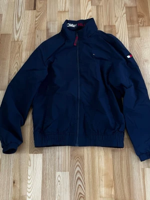 Marinblå vindjacka Tommy Hilfiger - Snygg marinblå vindjacka från Tommy Hilfiger med dragkedja framtill och diskret logga på bröstet samt ärmen. Jackan har hög krage, elastiska muddar och meshfoder på insidan för extra komfort. Perfekt för blåsiga dagar.