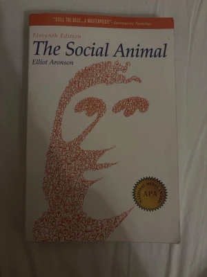 Bok: The Social Animal, Elliot Aronson - Kursbok på engelska: 'The Social Animal' av Elliot Aronson, elfte upplagan. Omslaget är vitt med en konstnärlig illustration i rött och blå text. Boken är inbunden och används ofta inom psykologi och samhällsvetenskap. Perfekt för dig som pluggar socialpsykologi.