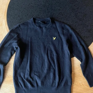 Svart stickad tröja från Lyle & Scott - Svart stickad tröja från Lyle & Scott med klassisk gul broderad fågel på bröstet. Tröjan har rund hals, långärmad passform och sitter snyggt med en normal fit. Perfekt för dig som gillar stilrena och enkla plagg med en ikonisk detalj.