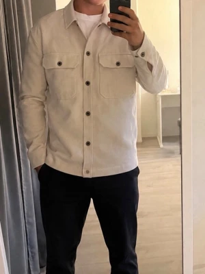 Beige overshirt med bröstfickor - Stilren beige overshirt med raka linjer och två stora bröstfickor med lock och knappar. Skjortan har klassisk krage och knäppning framtill med mörka knappar. Tillverkad i ett mjukt, tjockare tyg som ger en avslappnad men snygg look.