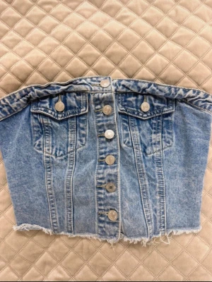 Ljusblå jeans tubtopp med knappar - Säljer en snygg ljusblå jeans tubtopp med silverfärgade knappar framtill och två dekorativa bröstfickor. Toppen har rå kant nertill för en trendig look och är perfekt för dig som vill sticka ut. Passar till både kjol och byxor.