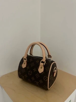 Brun monogram handväska med läderdetaljer - Snygg handväska i klassiskt brunt monogrammönster med ljusa läderhandtag och detaljer. Väskan har rundad form, guldfärgade metalldetaljer och dragkedja upptill. Perfekt storlek för det viktigaste och en ikonisk look.