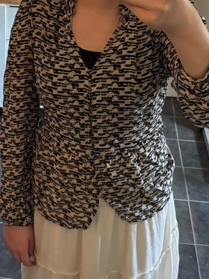 Mönstrad blus från InWear - Snygg blus från InWear i svartvitt abstrakt mönster. Blusen har v-ringning med liten volangkant och långa ärmar med resår vid handlederna. Luftig och lätt i materialet, perfekt för en trendig look. Storlek 38.