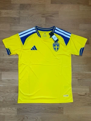 Svensk landslagströja Adidas gul XL - Gul svensk landslagströja från Adidas med blå detaljer och vita ränder på axlarna. Tröjan har broderat Sverige-emblem på bröstet och Adidas-logga i blått. Tillverkad i lätt och ventilerande material, perfekt för fotboll eller träning.