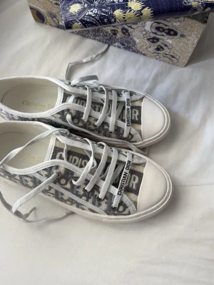 Christian Dior gråvita sneakers - Snygga sneakers från Christian Dior i grått och vitt med klassiskt Dior-mönster på canvas och vita gummisulor. Skorna har vita snören och detaljer med logga på plösen och sidan. Perfekta för dig som vill ha en exklusiv touch till din outfit.