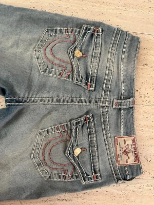 Ljusblå True Religion jeans med kontrastsömmar - Säljer ett par ljusblå jeans från True Religion med ikoniska kontrastsömmar och rosa detaljer på bakfickorna. 